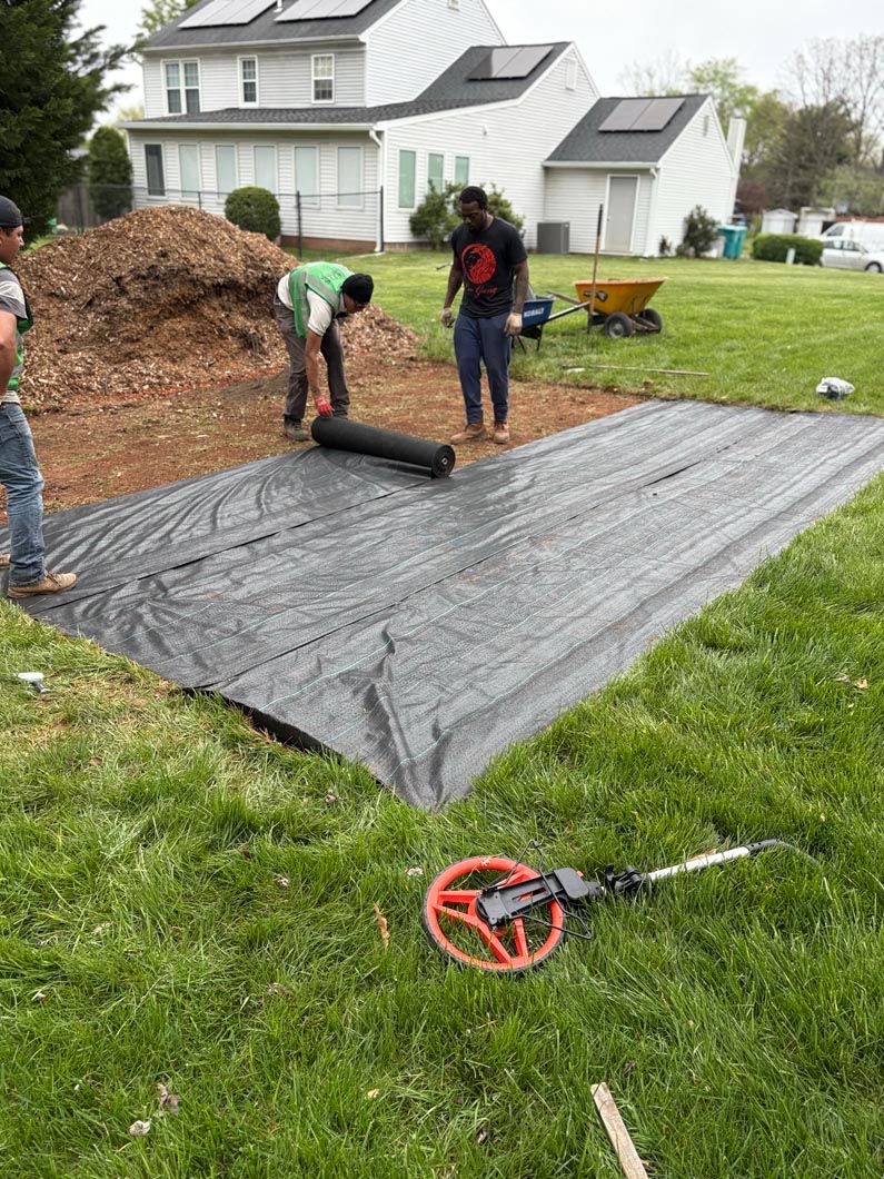 Workers laying black landscape fabric in a yard beside a mulch pile and house