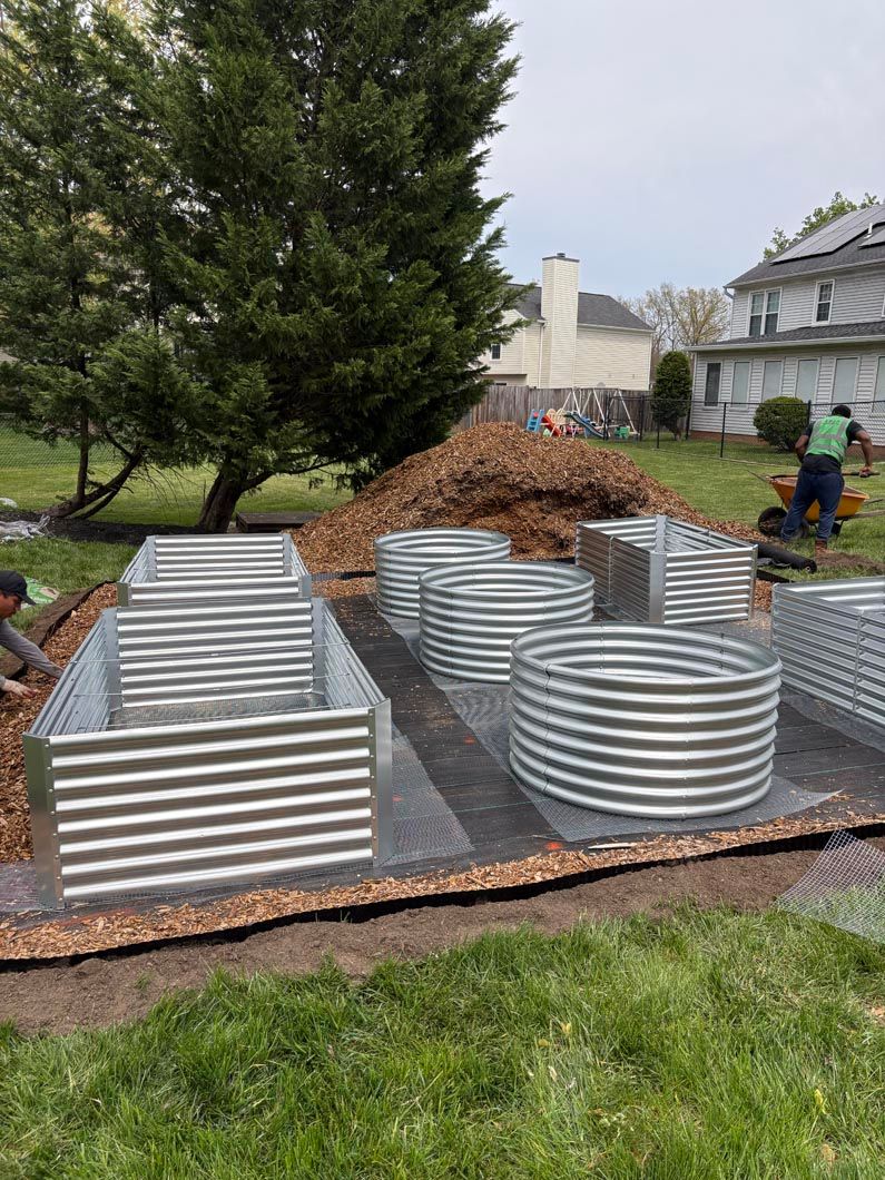 Workers assembling corrugated metal garden beds on black fabric in a yard beside a large tree.