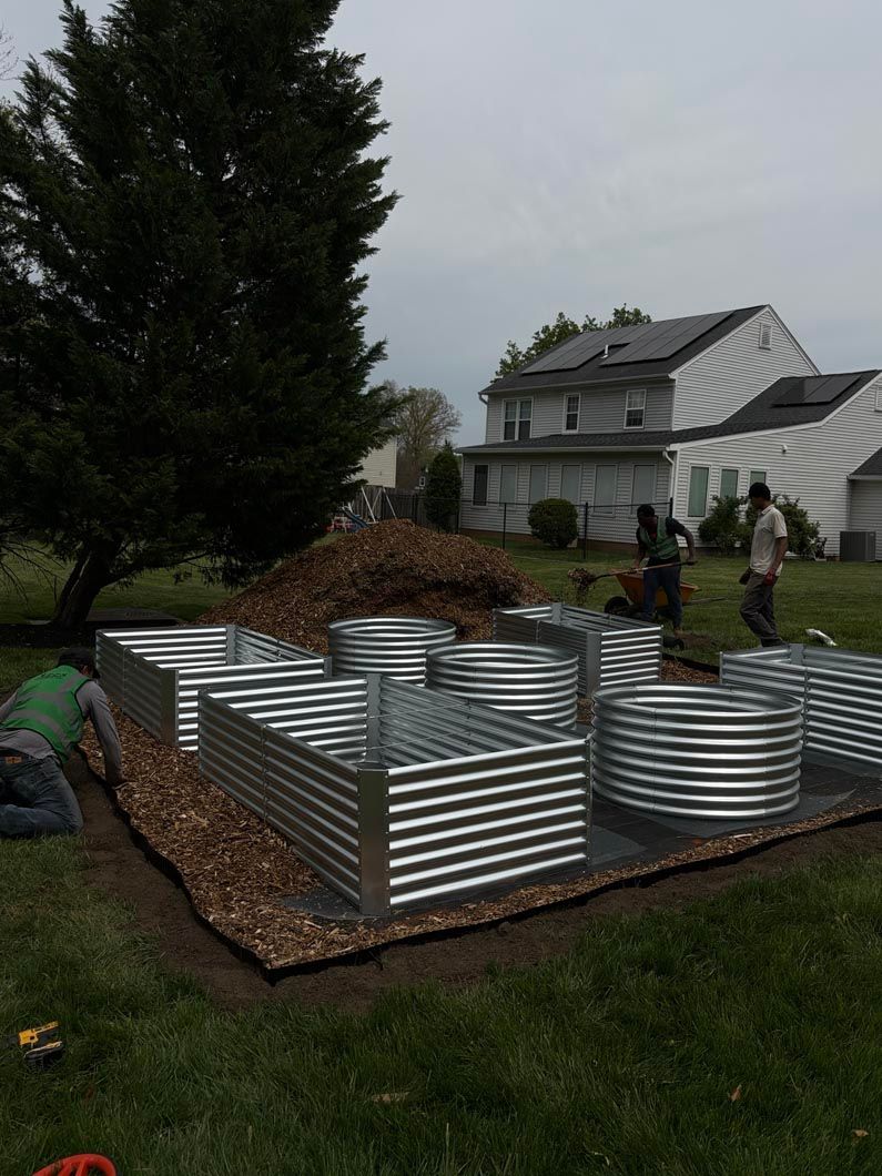 Raised corrugated metal garden beds in a yard beside a pile of leaves, with two people near a house.