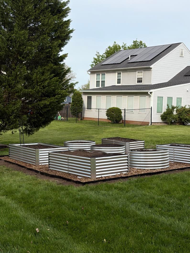 Raised corrugated metal garden beds in a grassy yard beside a white house and a large tree