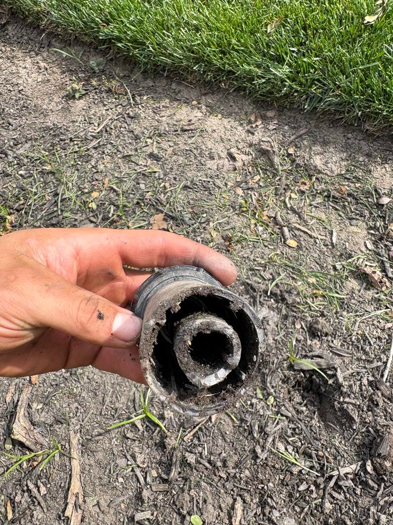 Hand holding a dirty, broken circular pipe fitting over muddy ground near grass