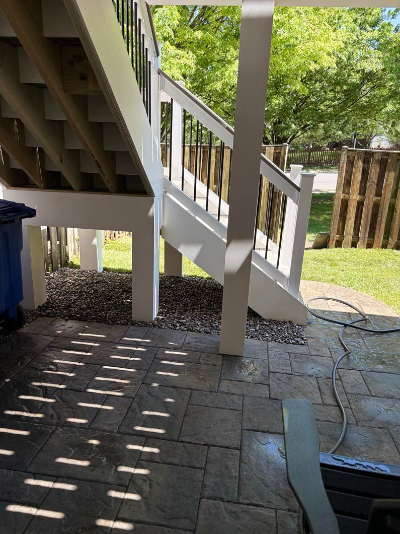 Covered patio with white stairs, concrete floor, and dappled sunlight under a deck