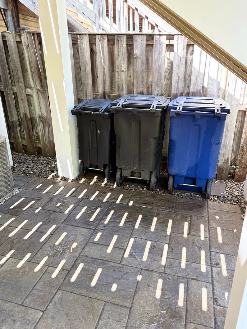 Three curbside trash bins, two black and one blue, beside a wooden fence on a paved patio.