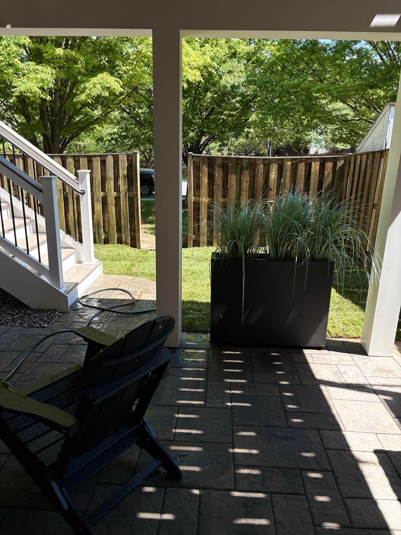 Shaded porch with a chair, potted plants, white stairs, and a wooden fence in the yard beyond