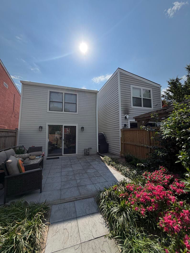 Backyard patio with white house, paved walkway, blooming pink flowers, and bright sun overhead