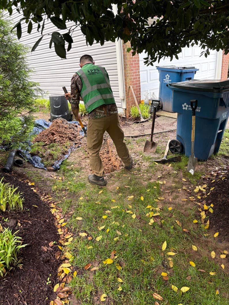 Person raking leaves in a backyard beside blue trash bins and a garden bed