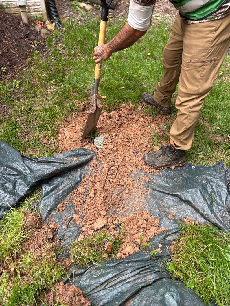 Person using a shovel to dig soil over black landscape fabric in a grassy garden area.