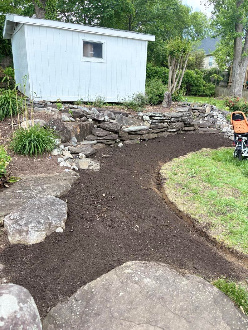 Freshly laid dark mulch path leading to a small white shed in a garden, with rocks and grass on either side.