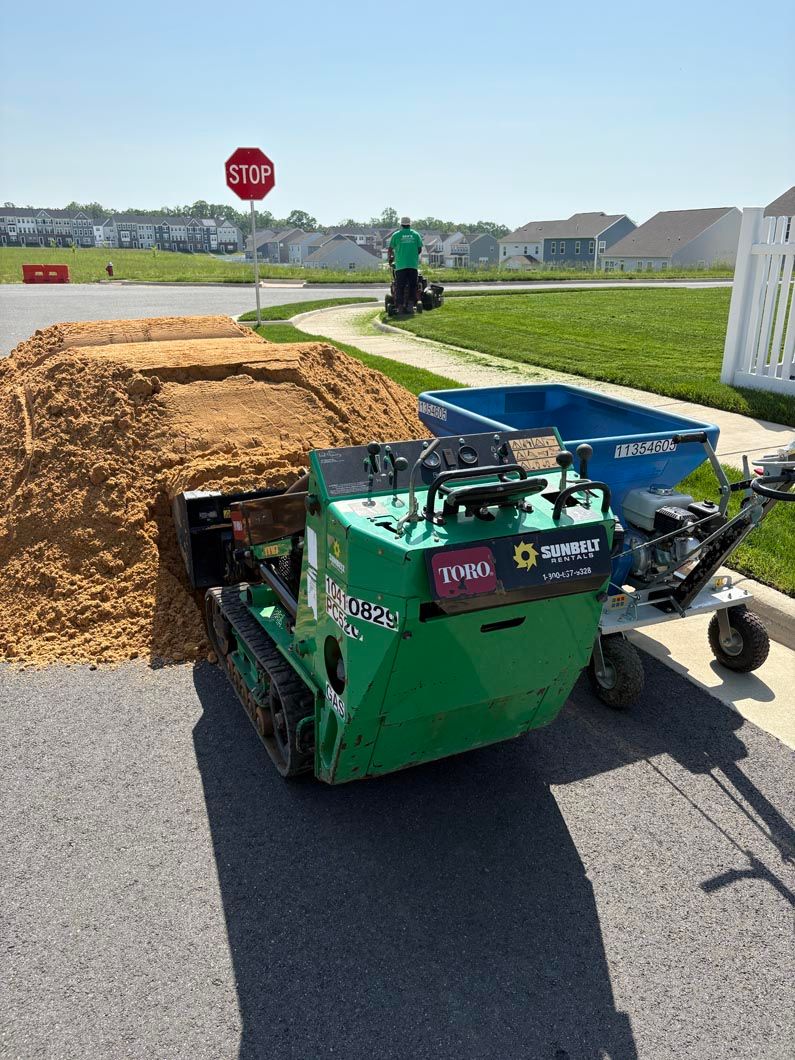 Green road roller beside a large asphalt pile at a street intersection with a stop sign and grassy lot.