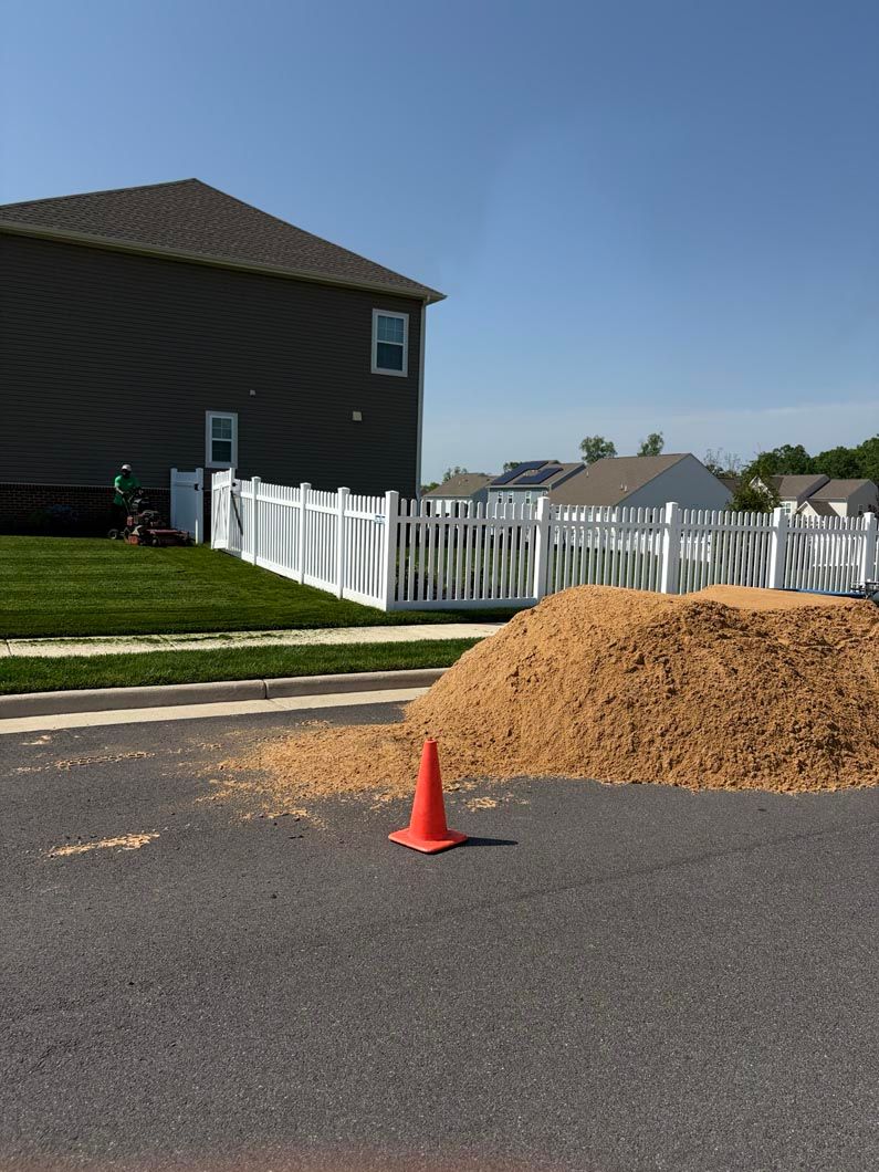 Orange traffic cone beside dirt pile on a residential street near a white fence and house