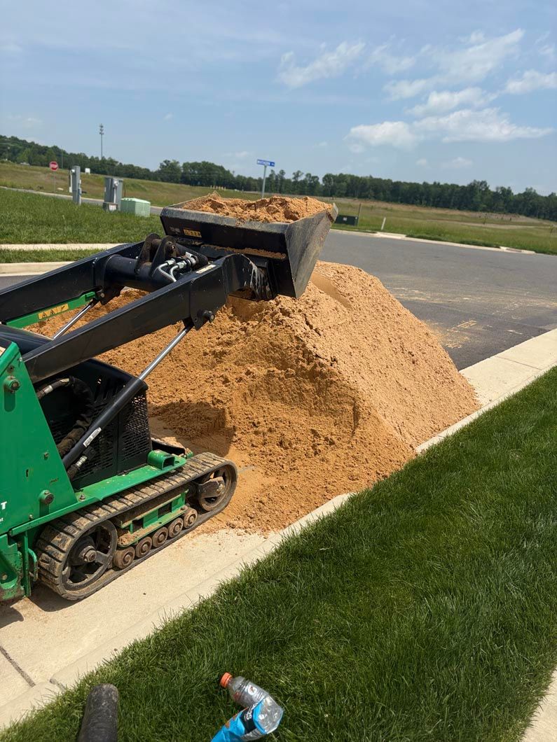 Green skid steer dumping a large pile of tan dirt beside a paved road and grassy field.