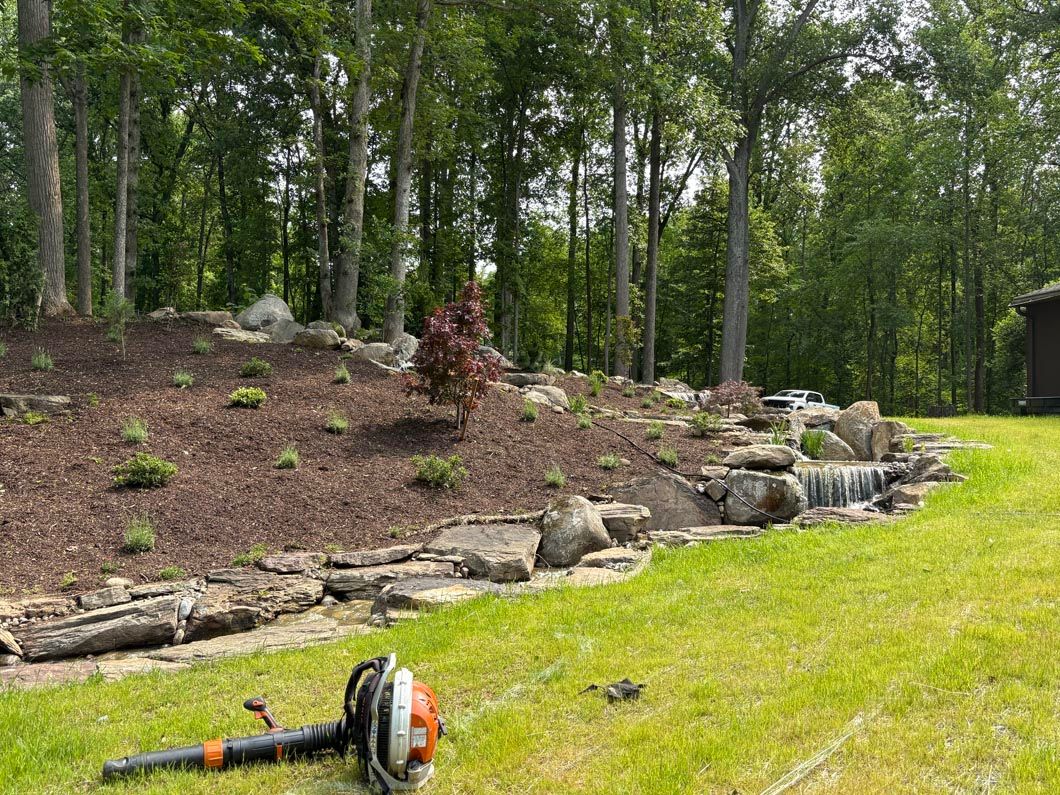 Garden bed with rocks and small shrubs beside a green lawn, with a leaf blower in the foreground