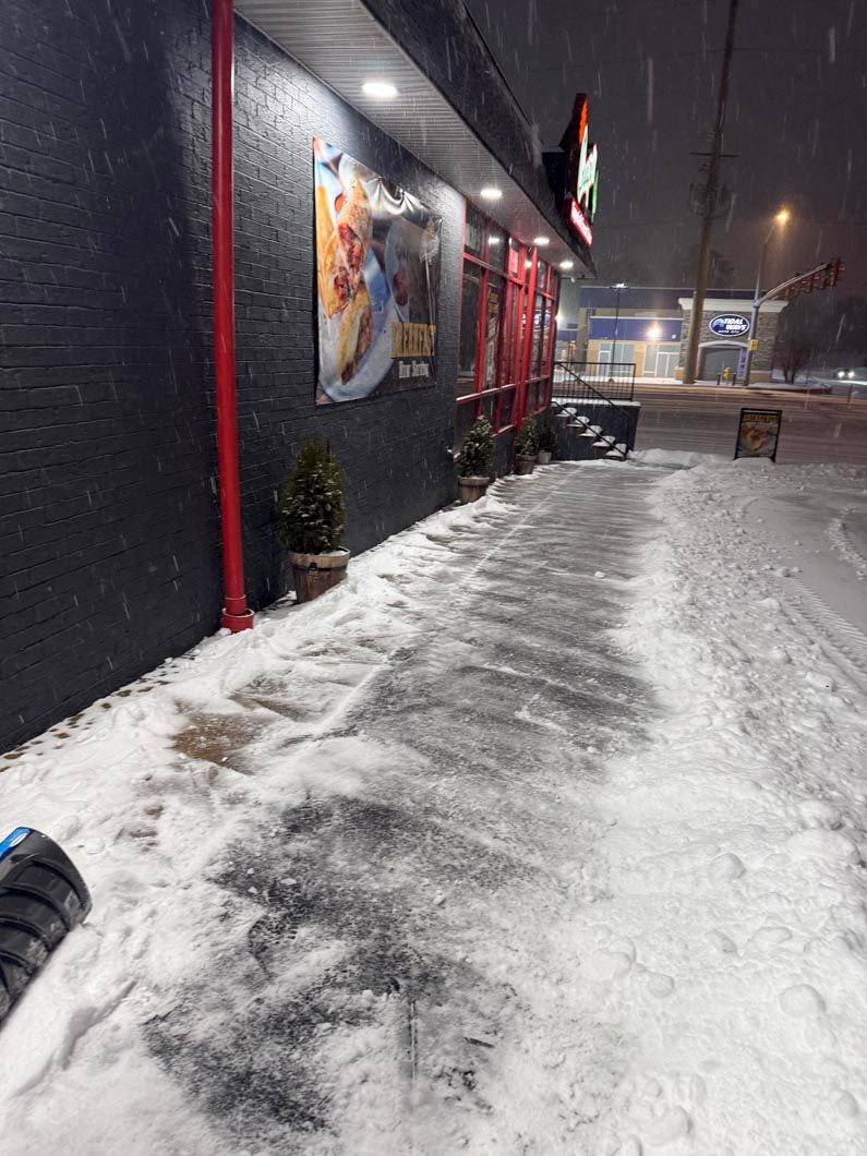 Snowy sidewalk outside a dark storefront at night, with red trim and falling snow.