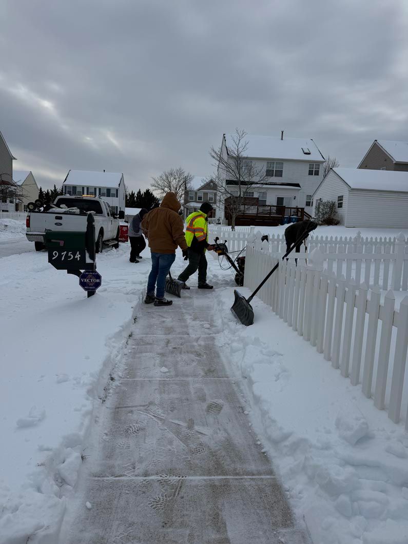 People shoveling a snowy sidewalk in a residential neighborhood, with a snow blower and parked trucks.