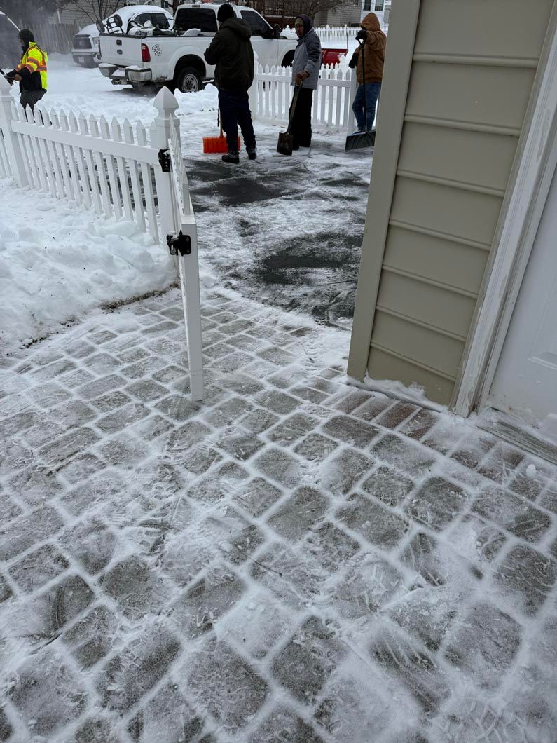 Snowy front walkway with shoveled path, white fence, and people standing in the yard