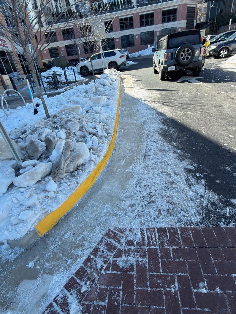 Snow-covered sidewalk with a yellow curb line beside a brick path and parked SUV.