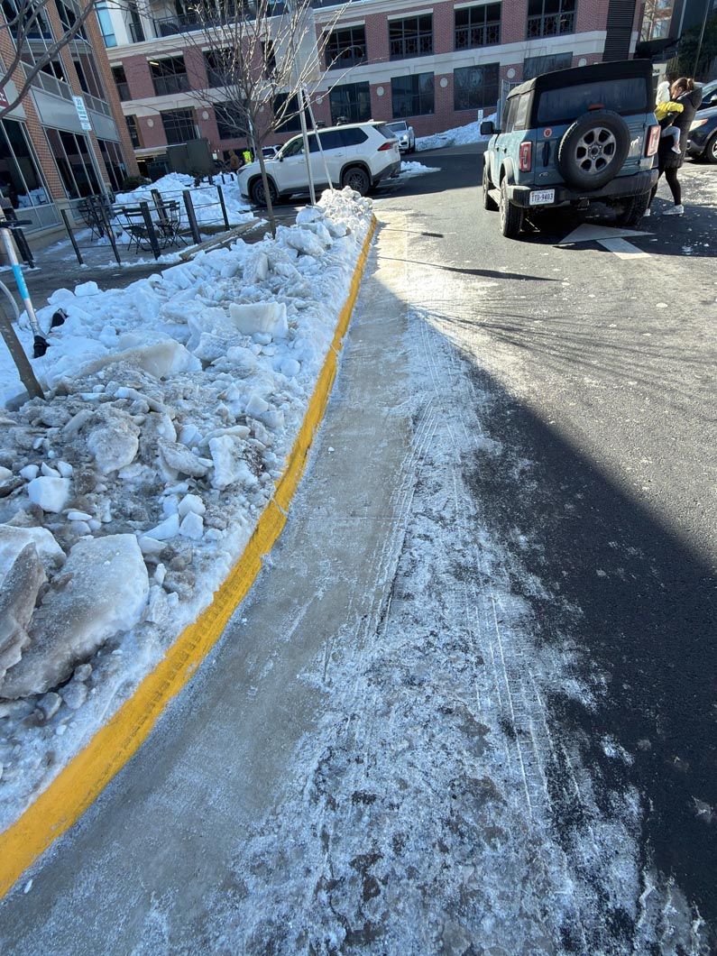 Snowy sloped sidewalk beside a brick building, with parked vehicles and a yellow curb line.