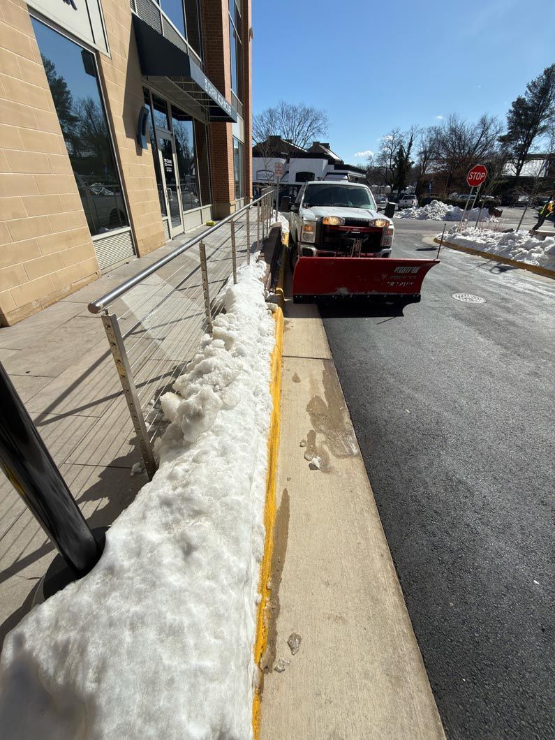 Red snowplow clearing snow along a curb beside a building on a snowy street.