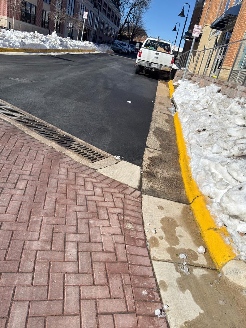 Snow-lined street with a white van parked by a yellow curb and brick sidewalk turning into asphalt