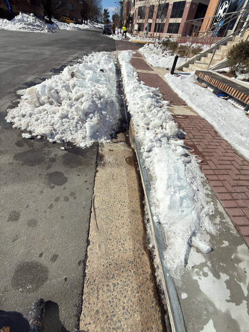 Snow piled along a sidewalk beside a road and building on a sunny day