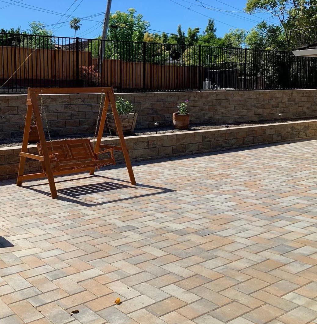 Wooden swing bench on a paved patio beside a stone wall and fenced garden under blue sky