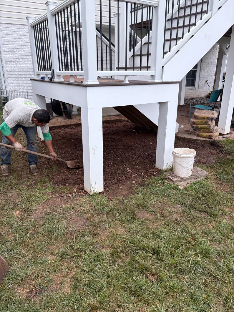 Person shoveling soil around a raised white deck with stairs and black railings in a yard