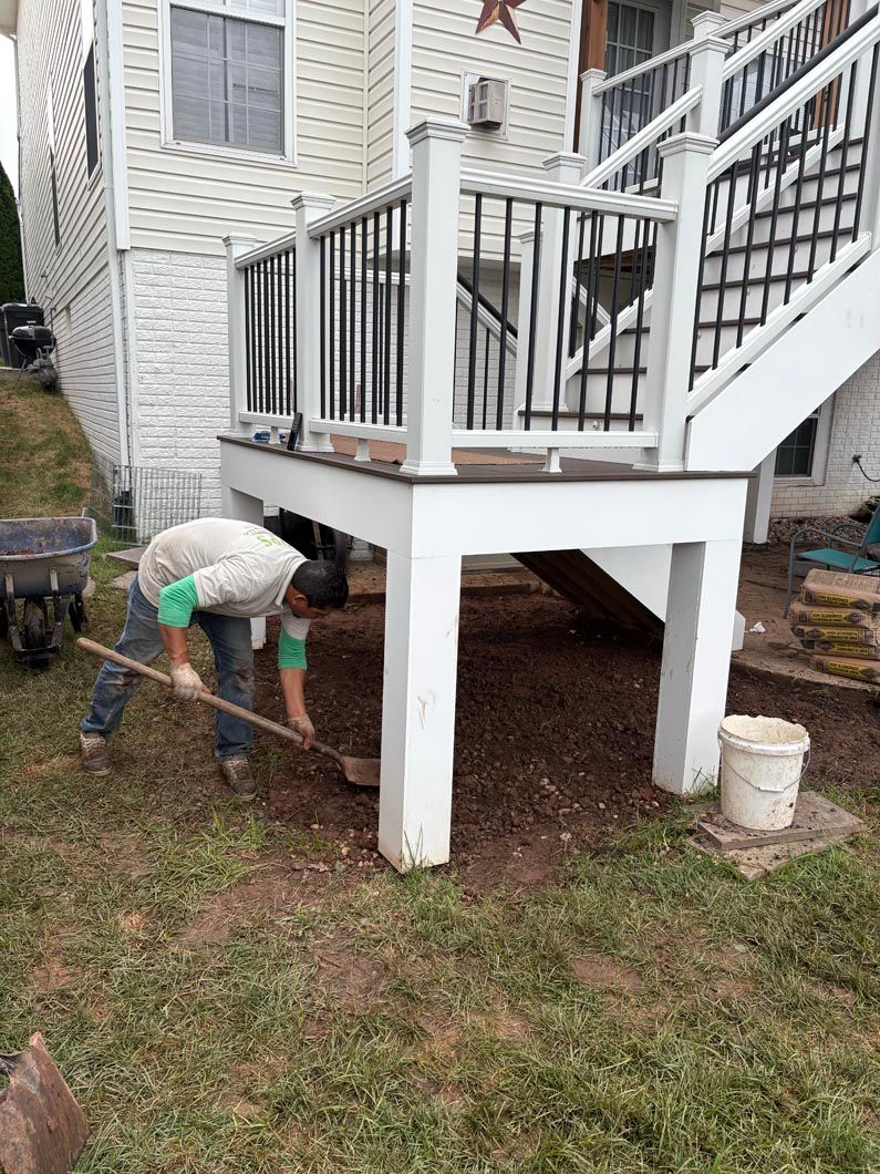 Person digging near a white house deck with railing, beside a bucket on the grass.