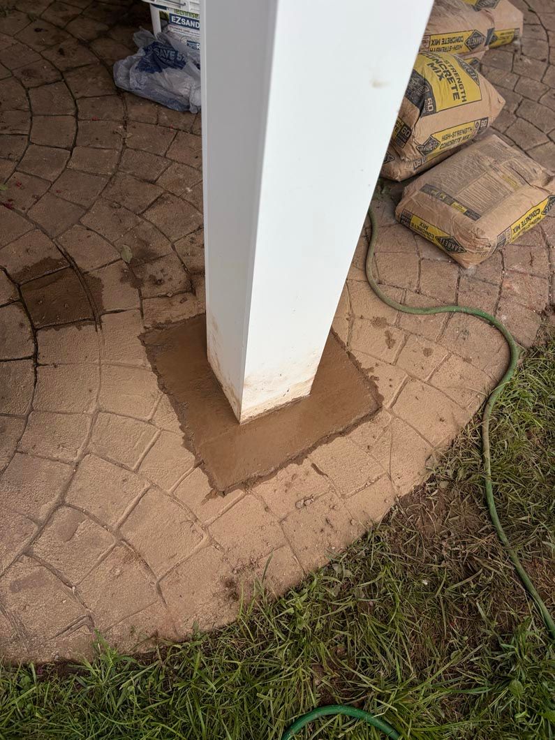 White pillar on tan paver patio with a brown repair patch at the base; grass and garden hose nearby