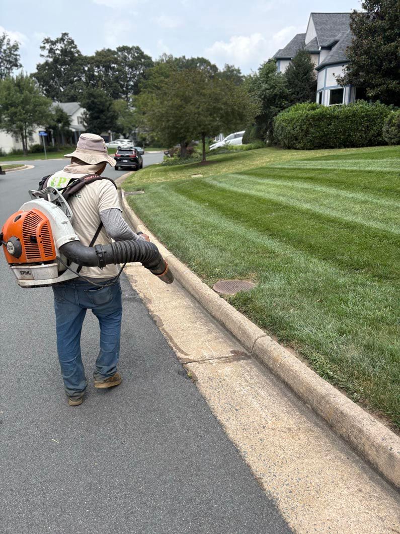 Worker in hat and backpack leaf blower cleaning a curb along a suburban street