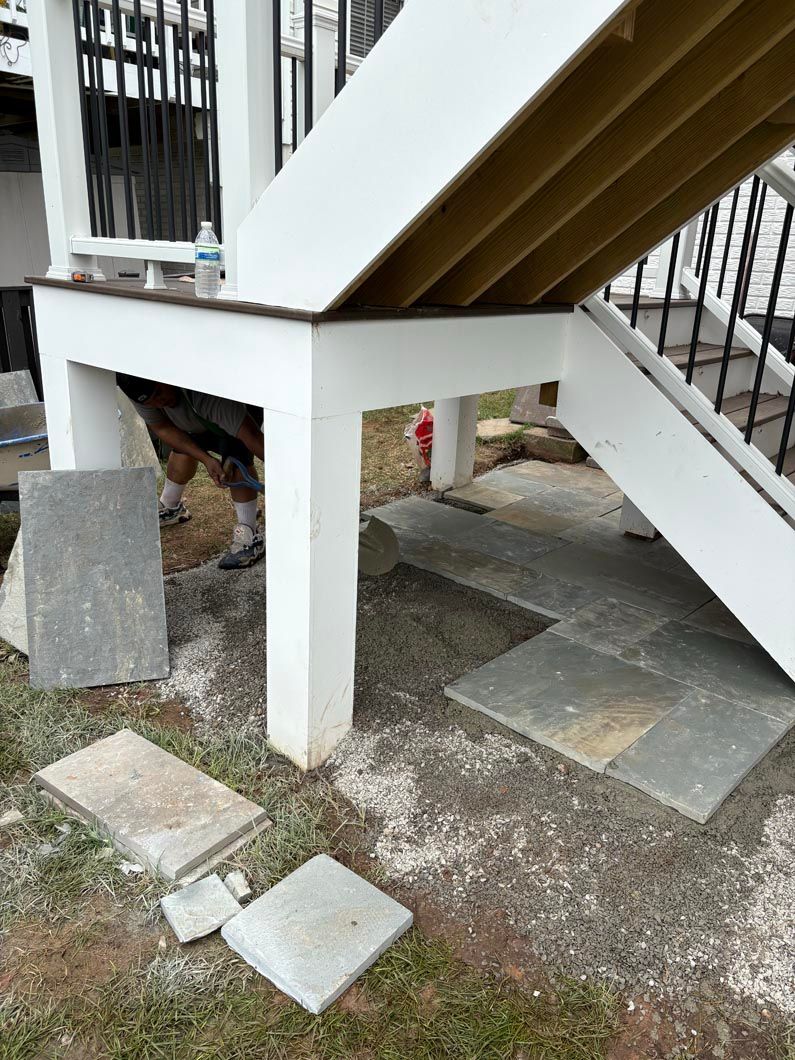 White staircase support pillar on uneven stone ground beneath an exterior stairway, with scattered concrete blocks.