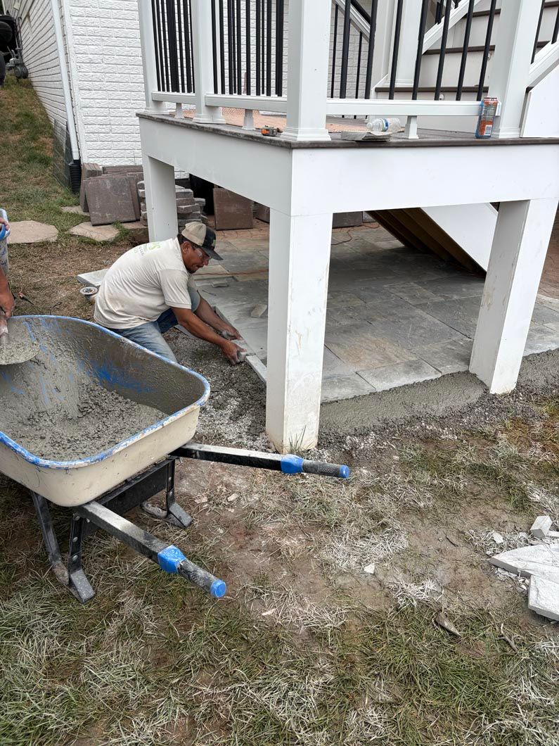 Worker painting the underside of a raised porch while crouching beside a wheelbarrow.