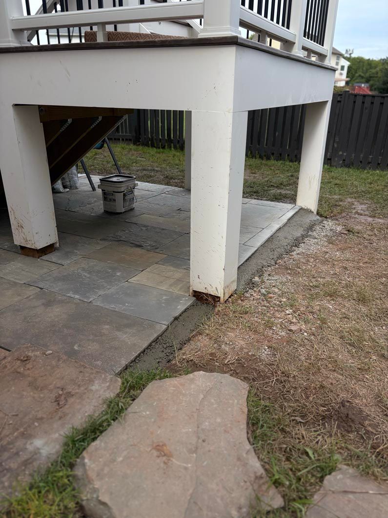 Concrete patio steps beside a raised deck, with a small bucket underneath and a large stepping stone in front.