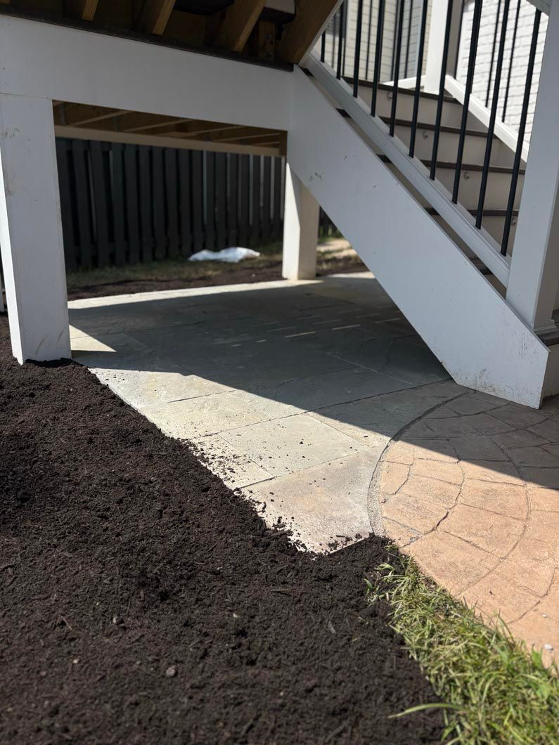 Concrete walkway under outdoor stairs with white railings and a small patch of grass nearby
