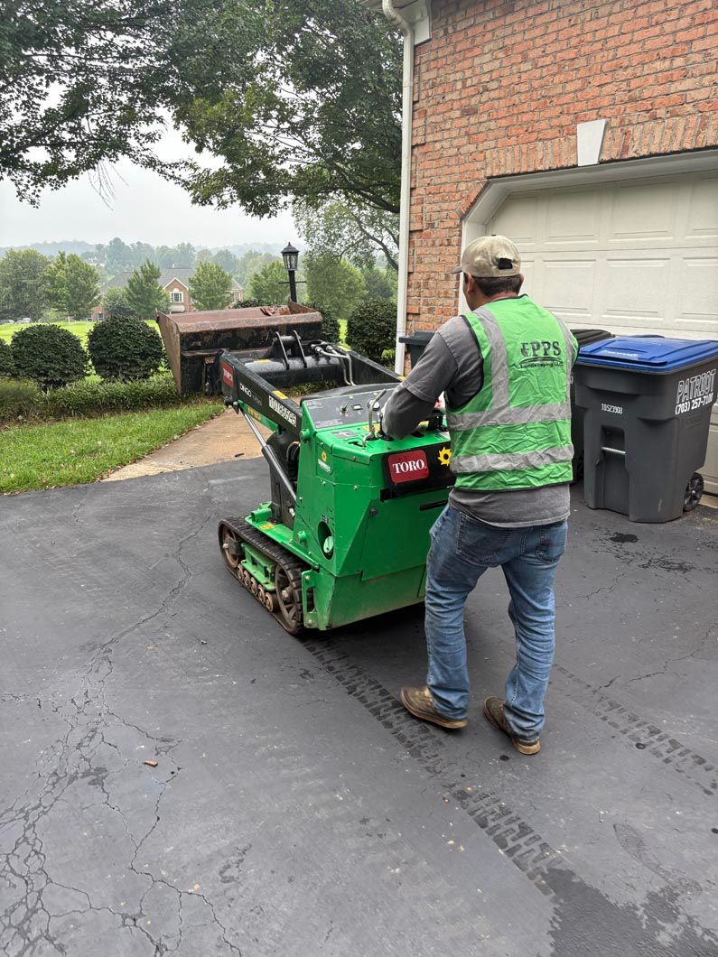 Worker operating a green skid steer on a driveway beside a brick garage and trash bins