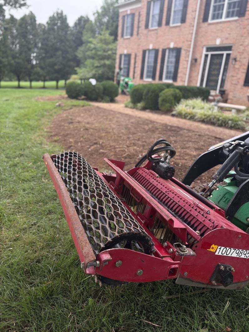 Red lawn aerator attached to a tractor beside a brick house and grassy yard.