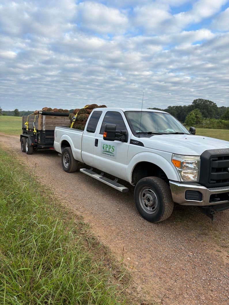 White utility truck hauling a trailer on a gravel road beside grassy fields under a cloudy sky