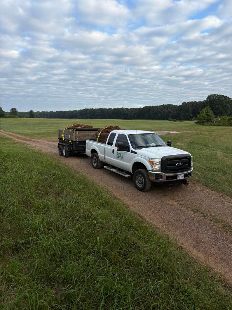 White pickup truck towing a flatbed trailer on a gravel road beside green fields under a cloudy sky