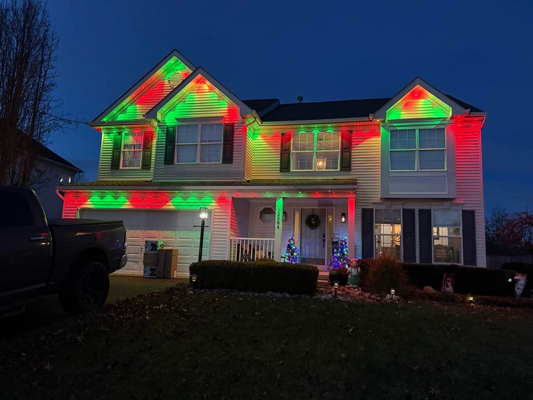 Two-story house at night lit with red and green holiday lights, with a dark truck parked beside it.