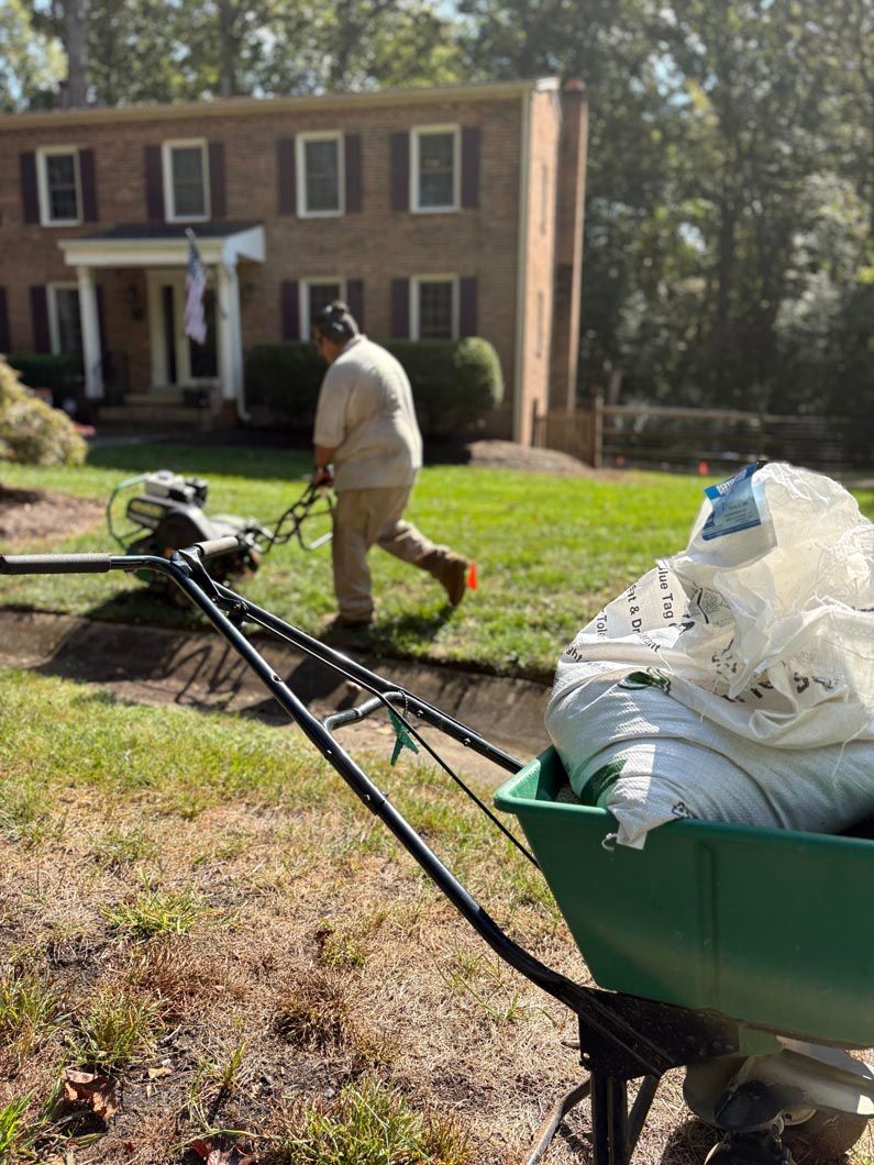 Person pushing a green yard cart across a grassy lawn in front of a brick house
