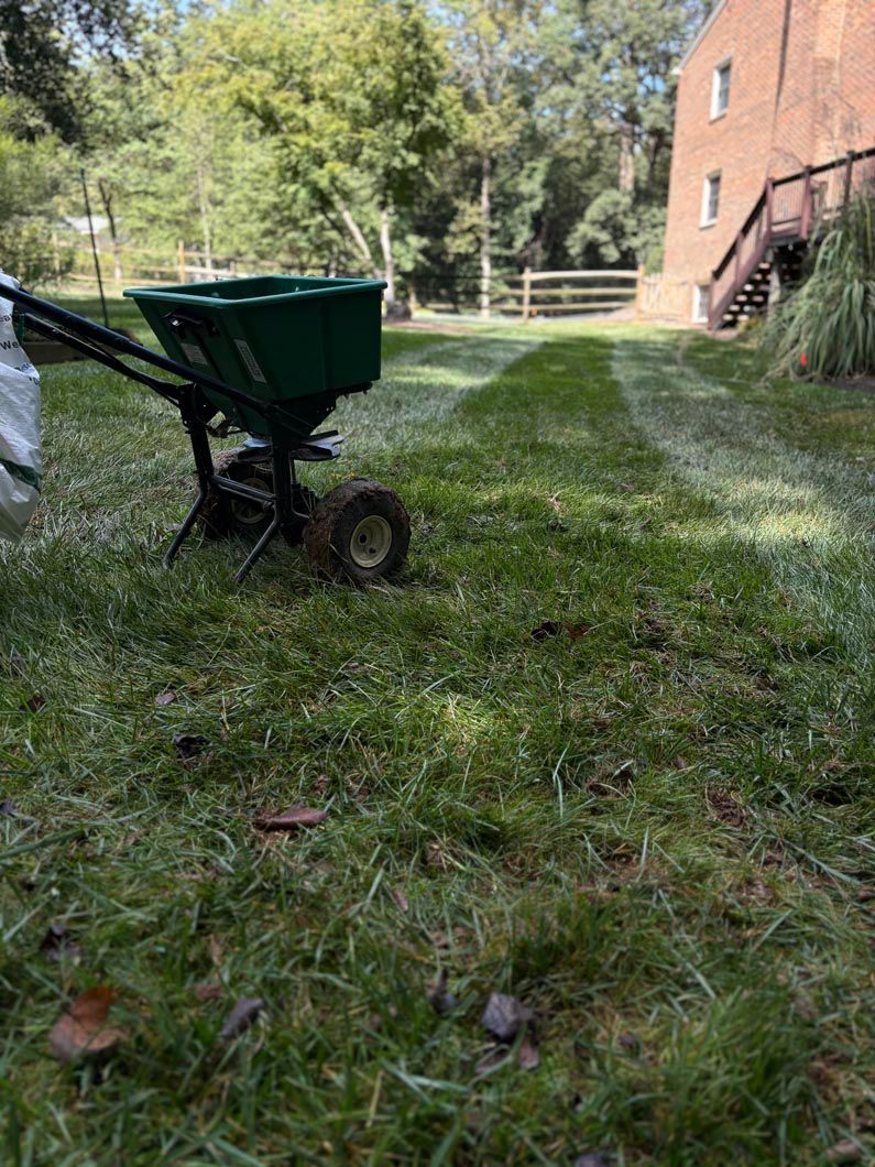 Wheelbarrow with green bin on a grassy yard beside a brick building and trees