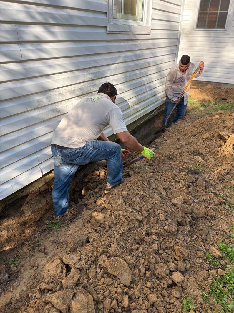 Two workers digging a trench along a white house’s foundation on a dirt slope