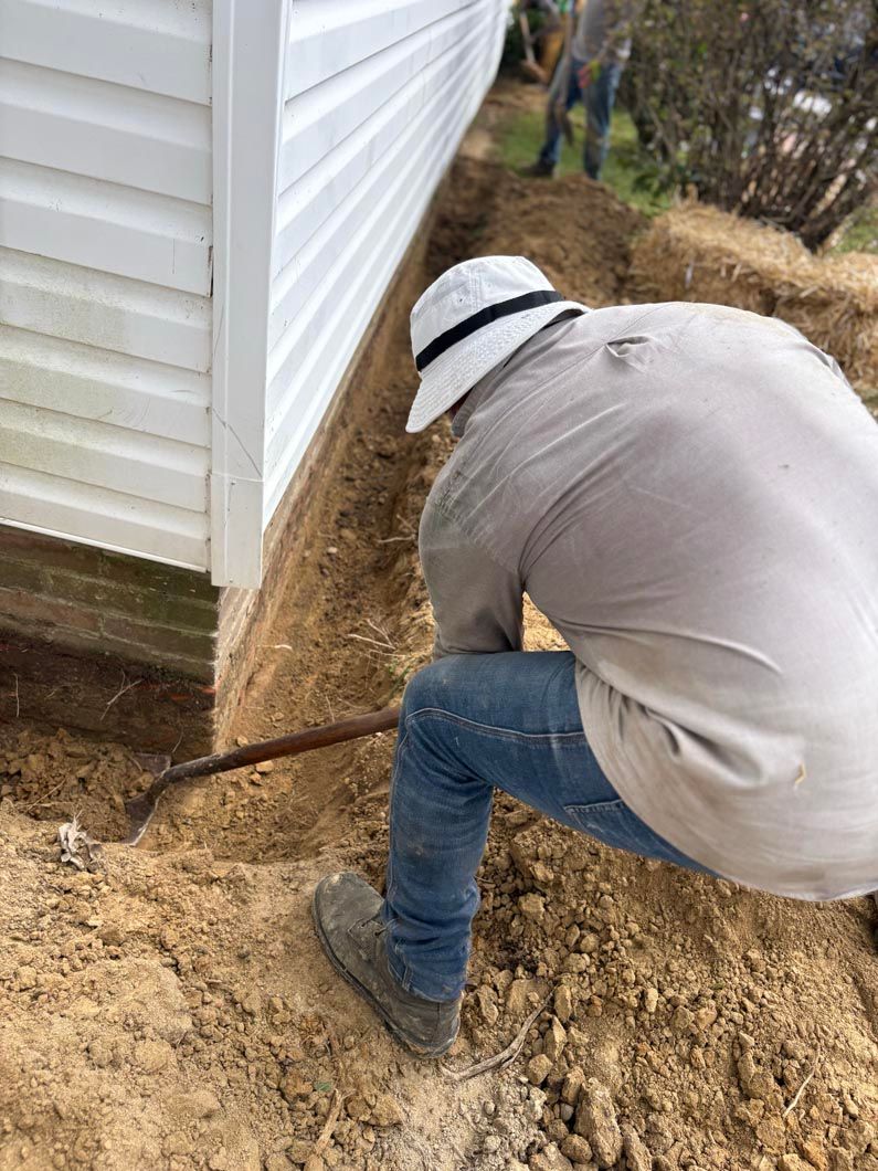 Person using a shovel beside a white house, digging along the foundation in muddy ground.