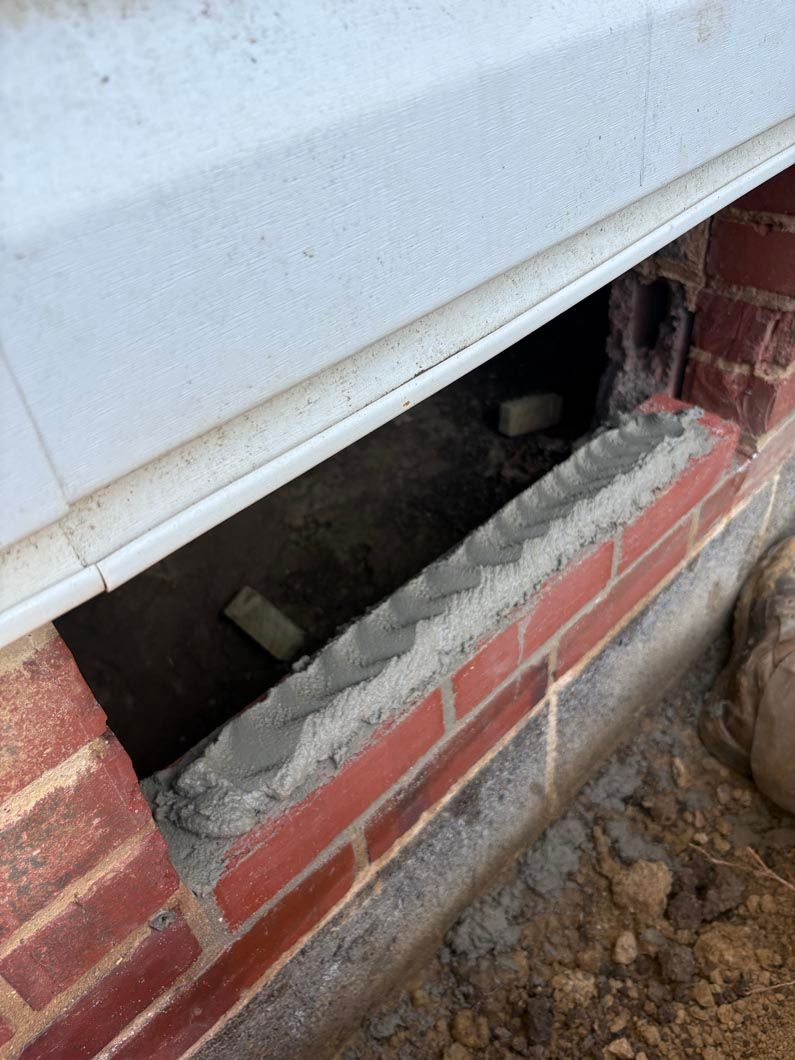 White siding lifted above a red brick foundation, exposing a dark gap with debris and dirt below