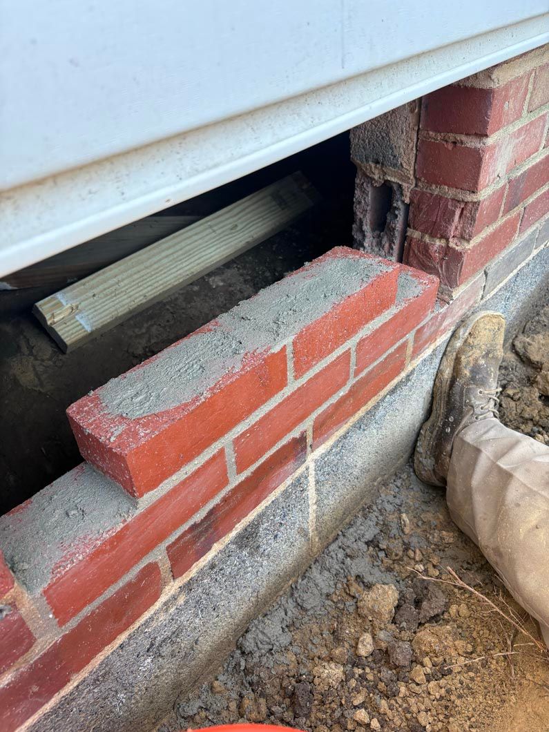 Broken red-brick wall beneath white siding, showing exposed gap and muddy ground below.