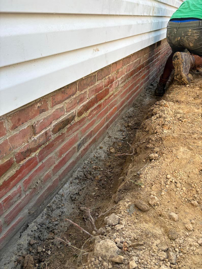 Foundation trench beside a house with white siding and brick veneer, showing exposed soil and excavation work