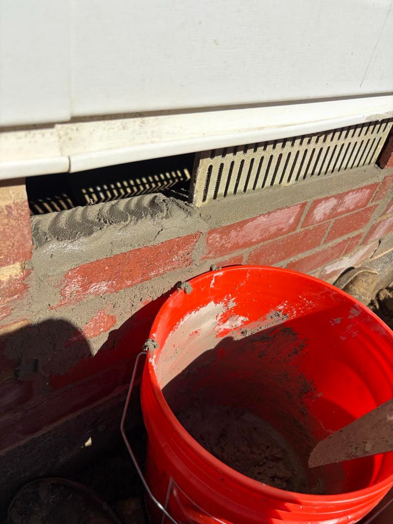 Red bucket beside a brick wall under a vent, with a concrete floor and debris.