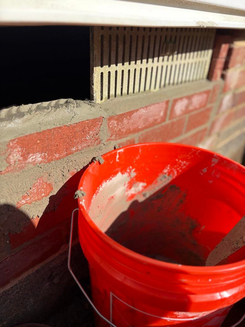 Red bucket with gray material on a ledge in front of a white fence.