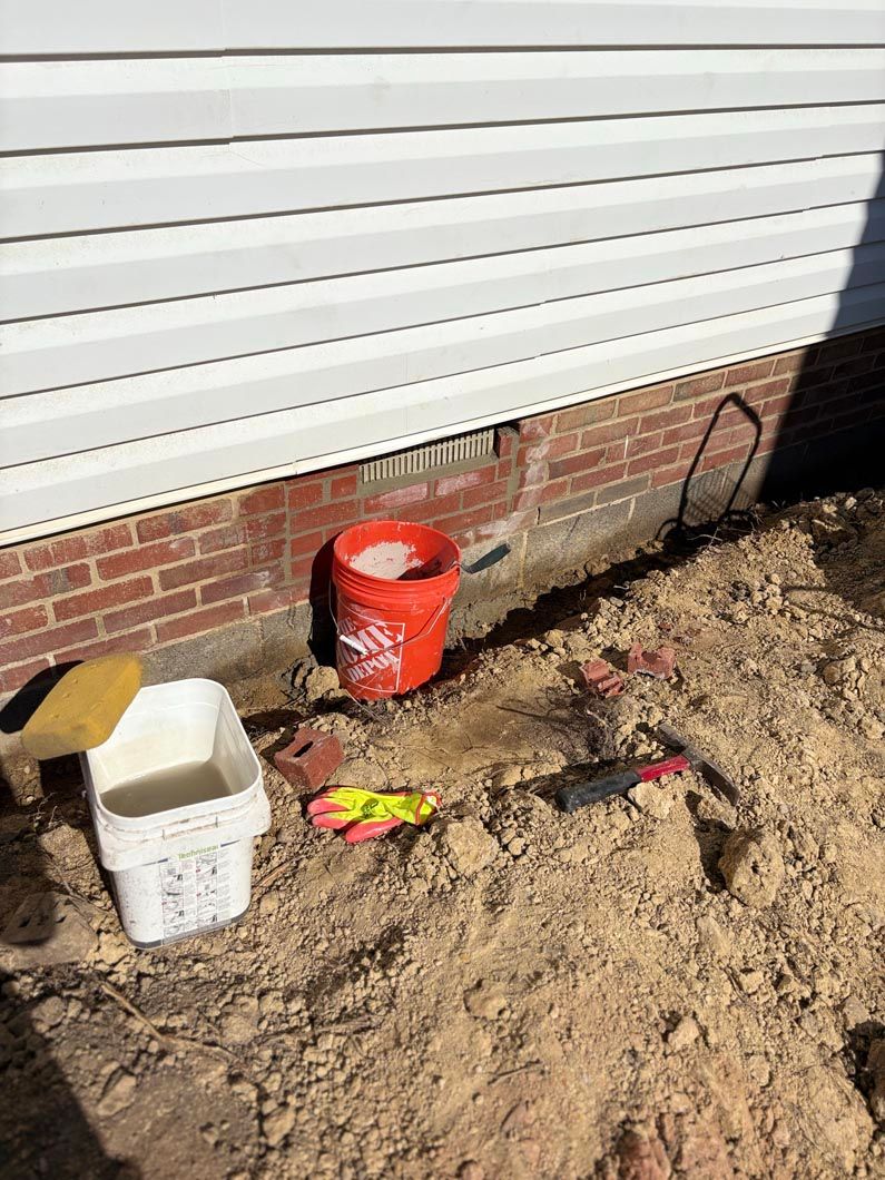 Construction materials and tools on dirt beside a brick house and white siding, including a red bucket and white bin
