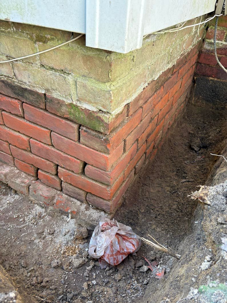 Brick foundation corner with damp soil and debris beside a white-sided building