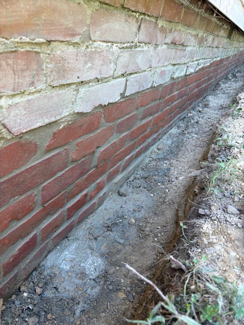 Brick wall with damaged plaster and a narrow trench along its base outdoors